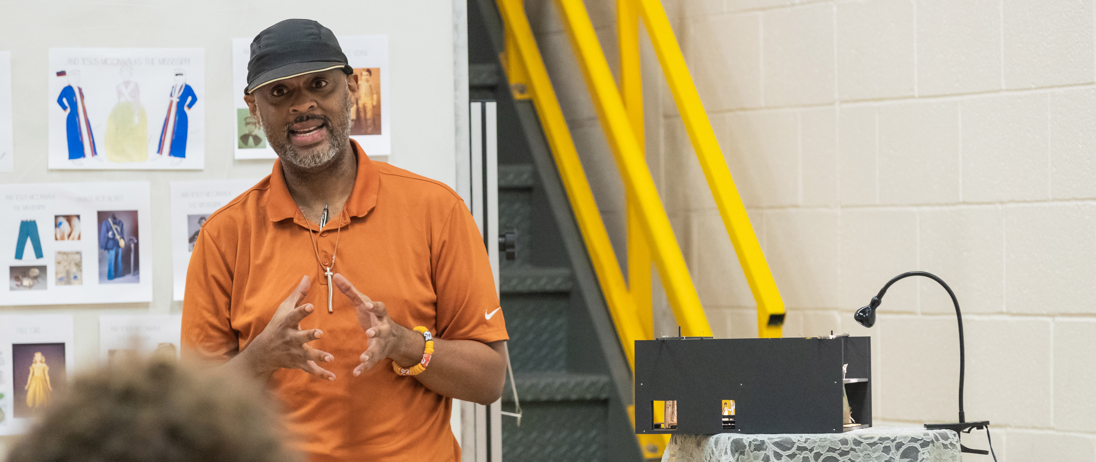 Director Christopher Burris addresses a group of students in the rehearsal hall. He stands in front of a bulletin board covered in costume design renderings. A small foam set model sets on a stool next to him.